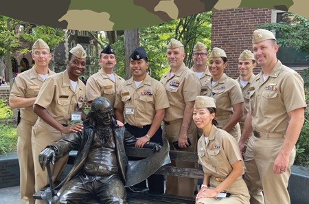Ten people in khaki U.S. Navy uniforms pose around a statue of Benjamin Franklin sitting on a bench.